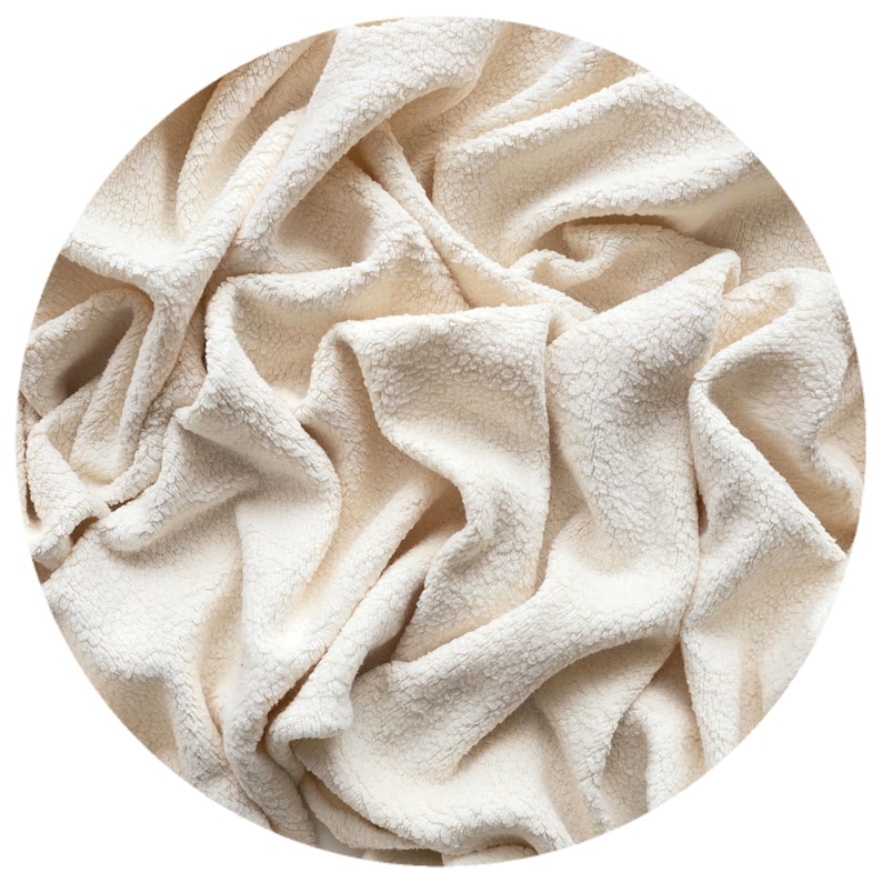 A close-up of a undyed natural cream-coloured, organic cotton sherpa fabric fleece with a textured, sherpa-like appearance. The fabric is heavily draped, creating deep folds and shadows. The organic cotton sherpa fabric appears soft and inviting.