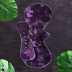 Puede incluir: Dos compresas menstruales de tela reutilizables en tonos morados con efecto tie-dye. Las compresas tienen forma contorneada con borde cosido blanco. El fondo es una superficie gris oscuro con hojas tropicales verdes.