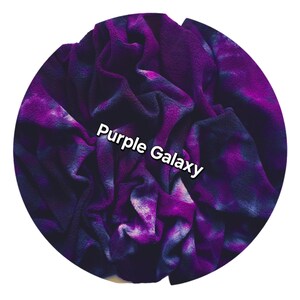 Organic cotton sherpa fabric with a tie-dye design in shades of purple and dark blue. The words "Purple Galaxy" are printed across the center in white. The blanket appears to be made of a plush material, ideal for comfort.