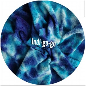 A circular image featuring a indigo blue and white tie-dye organic cotton sherpa fleece fabric swatch. The cotton sherpa fabric has a soft, textured appearance with indigo, turquoise, and white hues. The text "Indi-go-go" is displayed in the swatches.