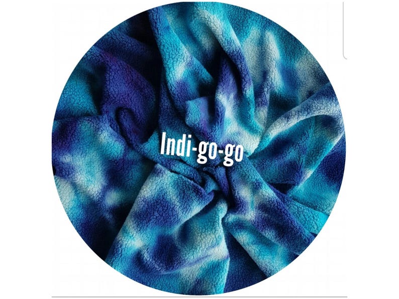 A circular image featuring a indigo blue and white tie-dye organic cotton sherpa fleece fabric swatch. The cotton sherpa fabric has a soft, textured appearance with indigo, turquoise, and white hues. The text "Indi-go-go" is displayed in the swatches.