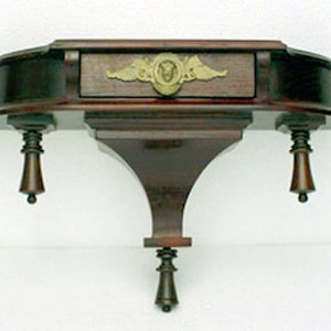 Puede incluir: Una estantería de pared de madera oscura con la parte superior curvada y un solo cajón. La estantería tiene detalles decorativos en la parte delantera y los lados, y el cajón tiene un tirador de latón con un diseño decorativo. La estantería está montada en la pared con dos soportes.