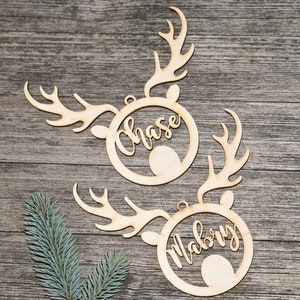 May include: Two wooden reindeer head ornaments with the names "Chase" and "Mabry" cut out in cursive script. The ornaments have antlers and a circular frame around the name. The background is a rustic wooden surface.