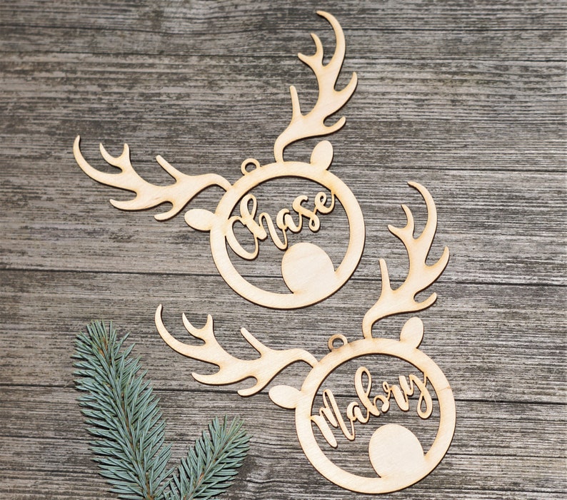 May include: Two wooden reindeer head ornaments with the names "Chase" and "Mabry" cut out in cursive script. The ornaments have antlers and a circular frame around the name. The background is a rustic wooden surface.