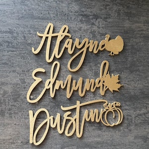 May include: Gold-colored wooden cutout names "Alayna", "Edmund", and "Dustin" with a turkey, maple leaf, and pumpkin cutout accents.
