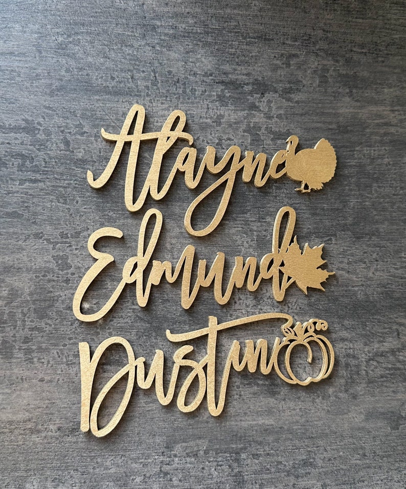May include: Gold-colored wooden cutout names "Alayna", "Edmund", and "Dustin" with a turkey, maple leaf, and pumpkin cutout accents.