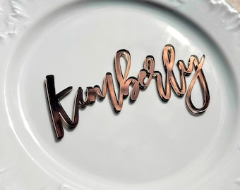 Puede incluir: Nombre "Kimberly" en acrílico oro rosa en fuente cursiva, colocado sobre un plato blanco con borde decorativo. El nombre es de un material brillante y reflectante, ideal para eventos.