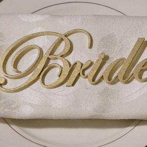 May include: A gold-colored "Bride" place card sits atop a folded white napkin on a white plate with a gold rim. The place card is in a cursive font with a textured surface, adding a touch of elegance to the table setting.