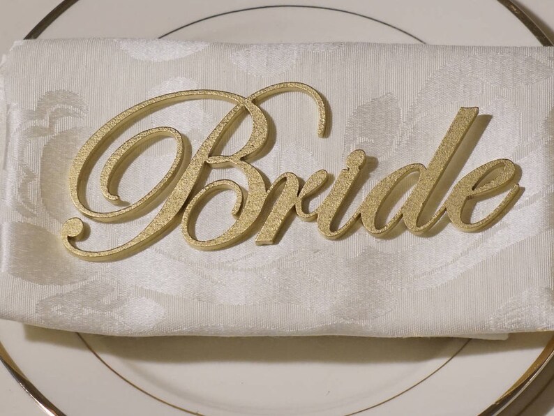 May include: A gold-colored "Bride" place card sits atop a folded white napkin on a white plate with a gold rim. The place card is in a cursive font with a textured surface, adding a touch of elegance to the table setting.