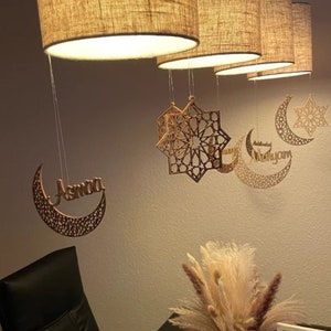 May include: Wooden crescent moon ornaments with intricate designs, including one with the name "Asmaa" and another with the word "Mubarakyum". The ornaments are hanging from light fixtures with fabric shades.