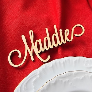 May include: Wooden name card with the name "Maddie" written in a cursive font. The card is on a red fabric background.