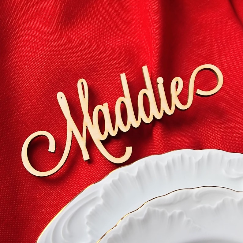 May include: Wooden name card with the name "Maddie" written in a cursive font. The card is on a red fabric background.