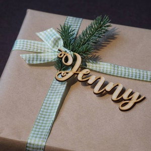 Puede incluir: Una bolsa de regalo de papel marr&oacute;n con una cinta de cuadros verdes y blancos y una etiqueta de madera con el nombre "Jenny".