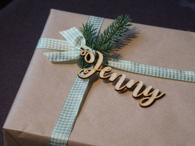Puede incluir: Una bolsa de regalo de papel marr&oacute;n con una cinta de cuadros verdes y blancos y una etiqueta de madera con el nombre "Jenny".