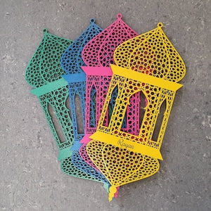 Puede incluir: Cuatro faroles de papel decorativos en diferentes colores, incluyendo amarillo, rosa, azul y verde. Los faroles están diseñados intrincadamente con patrones geométricos y tienen una forma recortada. El farol amarillo tiene la palabra "Rayan" escrita en él.