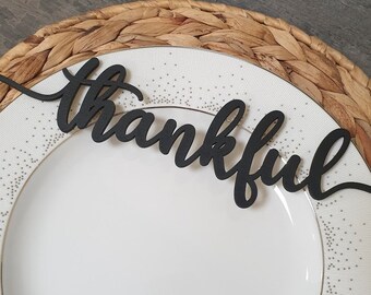 Könnte beinhalten: Ein weißer Teller mit Goldrand und dem dekorativen schwarzen Wort "thankful" in Schreibschrift liegt auf einem gewebten Platzset. Der Teller hat ein dezentes gepunktetes Muster um den Rand.