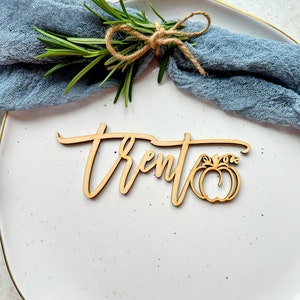 May include: Wooden place card with the name "Trent" written in cursive script and a small pumpkin cutout. The place card is on a white plate with a blue napkin and a sprig of rosemary.