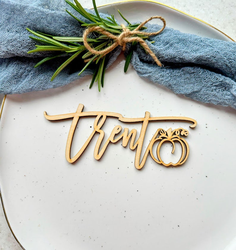 May include: Wooden place card with the name "Trent" written in cursive script and a small pumpkin cutout. The place card is on a white plate with a blue napkin and a sprig of rosemary.