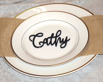 May include: A white plate with a gold rim and a black acrylic name card that says "Cathy" on it.