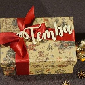 Puede incluir: Una caja de regalo marr&oacute;n con una cinta roja y una etiqueta de madera dorada que dice "Temba". La caja est&aacute; envuelta en papel con un dise&ntilde;o navide&ntilde;o vintage.