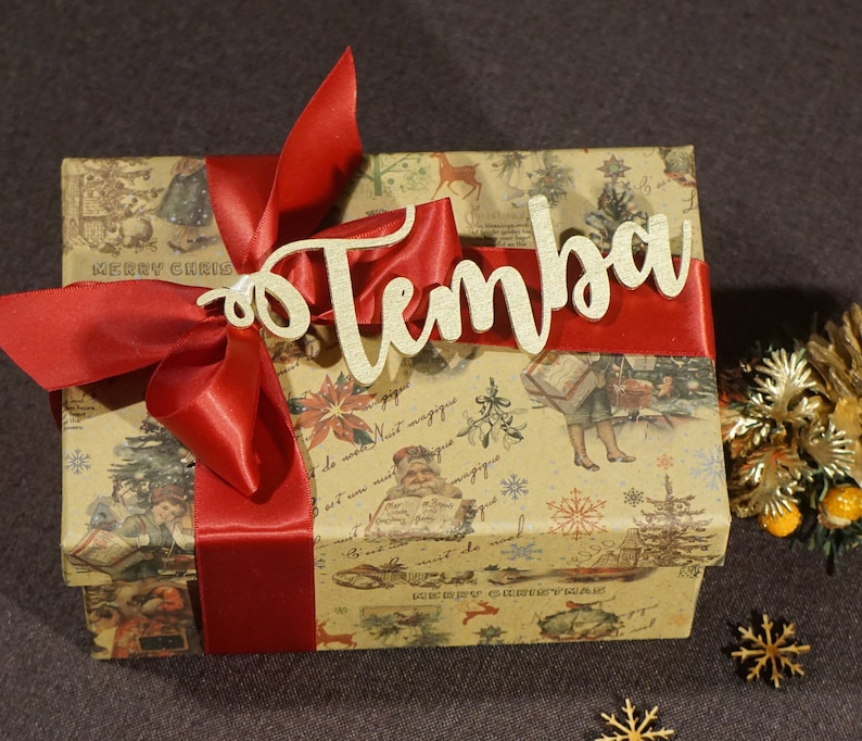 Puede incluir: Una caja de regalo marr&oacute;n con una cinta roja y una etiqueta de madera dorada que dice "Temba". La caja est&aacute; envuelta en papel con un dise&ntilde;o navide&ntilde;o vintage.