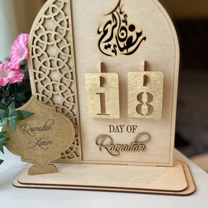 May include: A wooden Ramadan calendar with a gold-colored design. The calendar features a cutout of the Arabic word for Ramadan and a number display showing "18". The text "Day of Ramadan" is also displayed on the calendar.