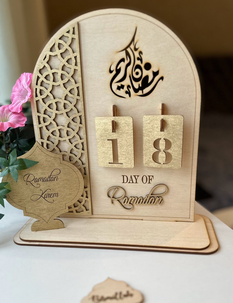 May include: A wooden Ramadan calendar with a gold-colored design. The calendar features a cutout of the Arabic word for Ramadan and a number display showing "18". The text "Day of Ramadan" is also displayed on the calendar.