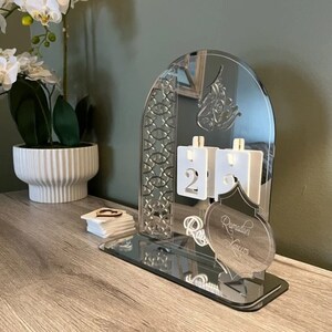 May include: A silver acrylic Ramadan countdown calendar with a mirrored base. The calendar features a decorative arch with an intricate design, a crescent moon, and the words "Ramadan Kareem".