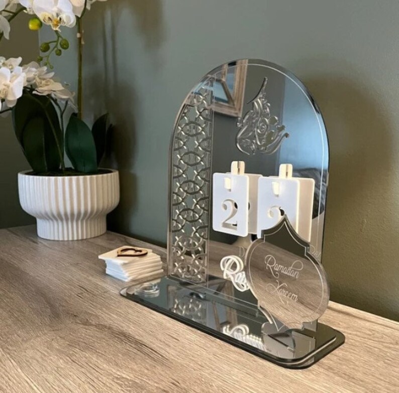 May include: A silver acrylic Ramadan countdown calendar with a mirrored base. The calendar features a decorative arch with an intricate design, a crescent moon, and the words "Ramadan Kareem".
