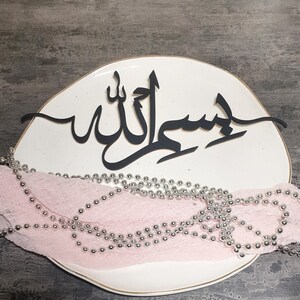 Op de afbeelding: Een wit keramiek bord met een zwart kalligrafieontwerp dat "Insha'Allah" zegt in Arabisch schrift. Het bord is versierd met een zilveren kralenkrans.