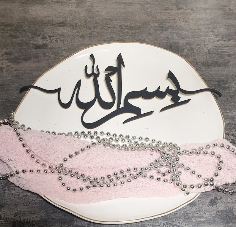 Op de afbeelding: Een wit keramiek bord met een zwart kalligrafieontwerp dat "Insha'Allah" zegt in Arabisch schrift. Het bord is versierd met een zilveren kralenkrans.