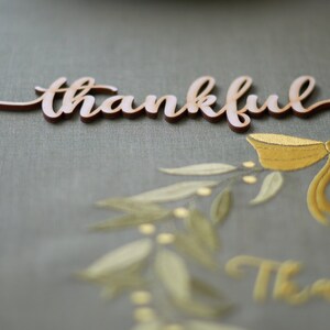 Pode incluir: Um recorte de madeira da palavra "thankful" em fonte cursiva, colocado acima de um desenho decorativo. O desenho inclui um la&ccedil;o dourado e elementos de ramo de oliveira, com a palavra "Thankful" repetida abaixo. O fundo &eacute; um tecido verde suave.