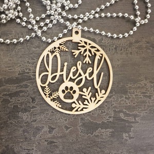 May include: A wooden Christmas ornament with the name "Diesel" cut out in a script font. The ornament is decorated with snowflakes and a paw print.