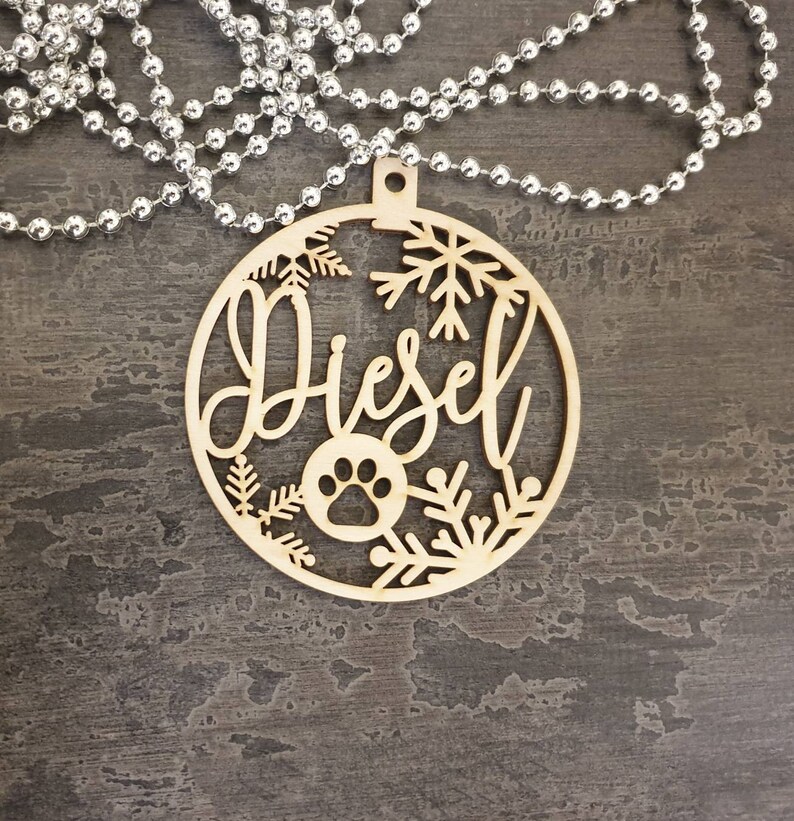 May include: A wooden Christmas ornament with the name "Diesel" cut out in a script font. The ornament is decorated with snowflakes and a paw print.