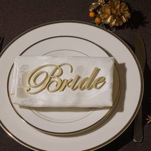May include: A gold-colored "Bride" place card rests on a folded white napkin, set on a white plate with a gold rim. A fork and knife are on either side. Decorative elements include wooden snowflakes and a heart.