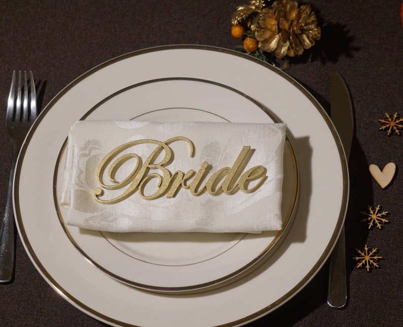 May include: A gold-colored "Bride" place card rests on a folded white napkin, set on a white plate with a gold rim. A fork and knife are on either side. Decorative elements include wooden snowflakes and a heart.