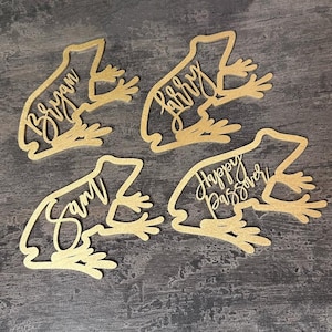 May include: Four laser-cut wooden frog-shaped place cards. Each frog has a name or phrase: "Bryan", "Larry", "Sam", and "Happy Passover" in a cursive font. The frogs are a light gold color and sit on a dark gray surface.