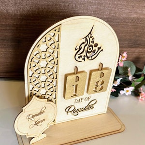 May include: Wooden Ramadan calendar with a cutout design. The calendar features a gold-colored background with the words "Day of Ramadan" and the numbers "18". The calendar also includes a small sign that says "Ramadan Kareem".