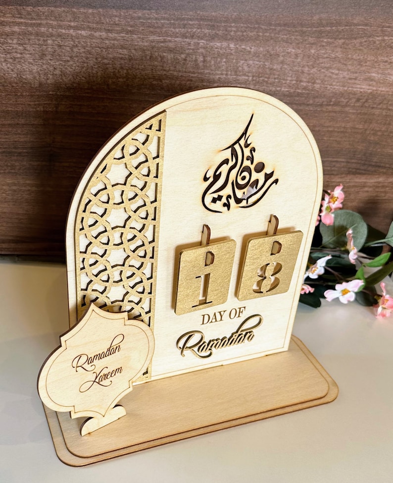 May include: Wooden Ramadan calendar with a cutout design. The calendar features a gold-colored background with the words "Day of Ramadan" and the numbers "18". The calendar also includes a small sign that says "Ramadan Kareem".