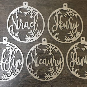 May include: Five white wooden ornaments with snowflake designs and personalized names:  "Nirael", "Heury", "Rafelin", "Nicoury", and "Daniel".