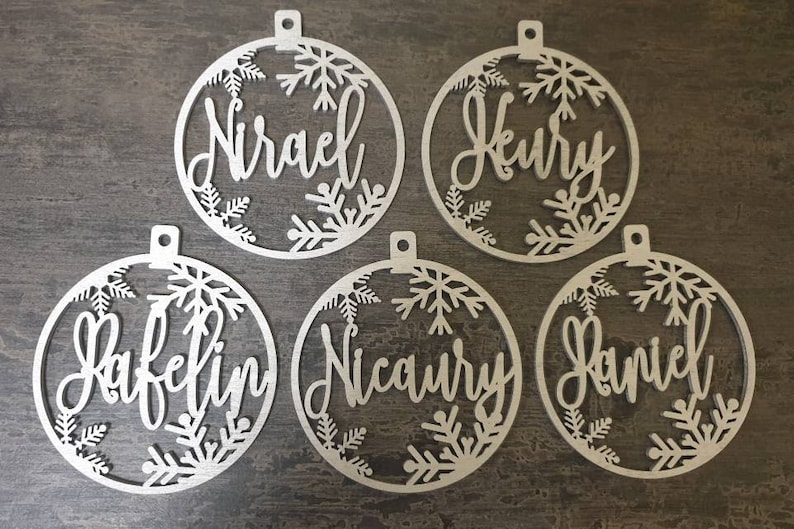 May include: Five white wooden ornaments with snowflake designs and personalized names:  "Nirael", "Heury", "Rafelin", "Nicoury", and "Daniel".