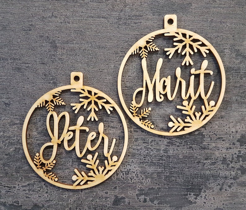 May include: Two wooden Christmas baubles, each with a name cut out in a cursive font: "Peter" and "Marit". The circular baubles feature snowflake designs around the edges and a small hole at the top for hanging, made from natural wood.