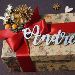 Puede incluir: Una cinta roja atada alrededor de una caja de regalo marr&oacute;n con un dise&ntilde;o navide&ntilde;o. La caja est&aacute; decorada con una pi&ntilde;a dorada y una etiqueta de nombre plateada que dice "Andr&eacute;".