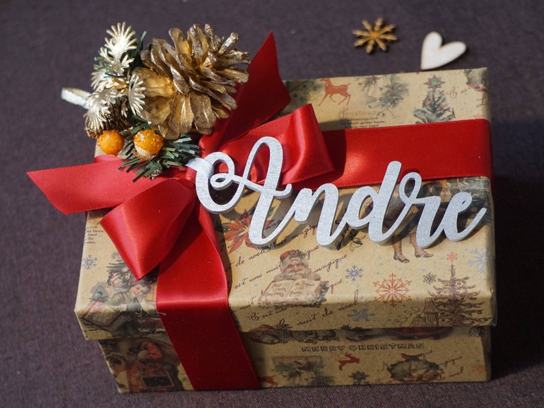 Puede incluir: Una cinta roja atada alrededor de una caja de regalo marr&oacute;n con un dise&ntilde;o navide&ntilde;o. La caja est&aacute; decorada con una pi&ntilde;a dorada y una etiqueta de nombre plateada que dice "Andr&eacute;".