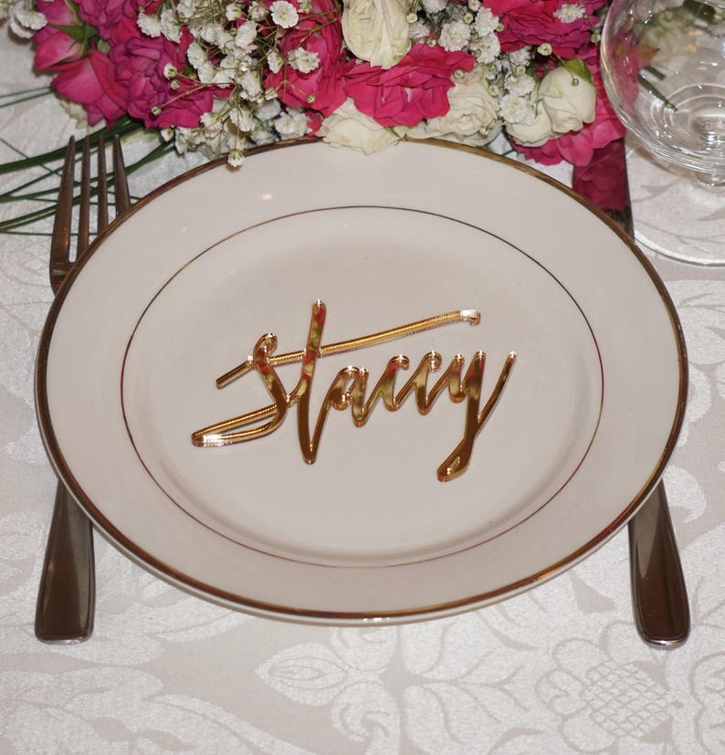 May include: A white plate with a gold rim and a gold acrylic name card that says "Stacy" on it. The plate is on a white tablecloth.