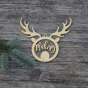 May include: A wooden Christmas ornament in the shape of a reindeer head with antlers. The name "Mabry" is written in cursive within the reindeer's face. The ornament is made of light-colored wood and has a small hole for hanging.