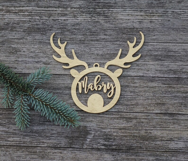 May include: A wooden Christmas ornament in the shape of a reindeer head with antlers. The name "Mabry" is written in cursive within the reindeer's face. The ornament is made of light-colored wood and has a small hole for hanging.