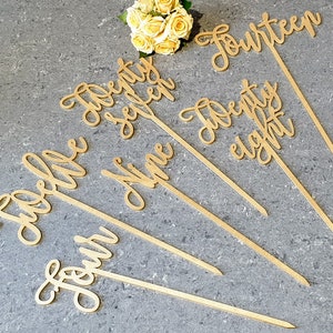 May include: Gold wooden table numbers on sticks, with the numbers "Four", "Nine", "Twelve", "Twenty-seven", "Twenty-eight", "Fourteen".
