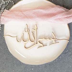 Op de afbeelding: Een houten bord met de Arabische woorden "Bismillah" en "Allah" in een decoratief schrift. Het bord ligt op een wit bord met een roze doek eroverheen gedrapeerd.