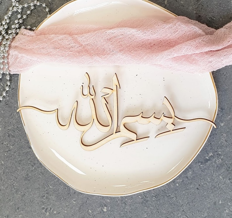 Op de afbeelding: Een houten bord met de Arabische woorden "Bismillah" en "Allah" in een decoratief schrift. Het bord ligt op een wit bord met een roze doek eroverheen gedrapeerd.